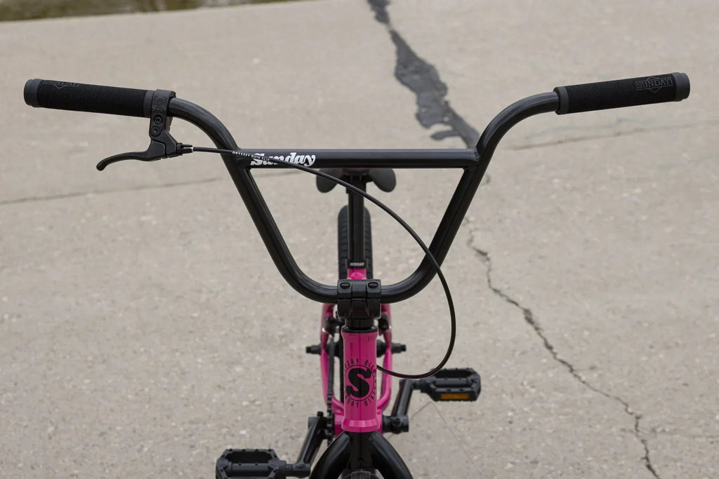 Vue rapprochée du guidon noir d'un vélo BMX avec cadre rose sur un sol en béton.
