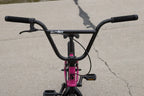 Vue rapprochée du guidon noir d'un vélo BMX avec cadre rose sur un sol en béton.