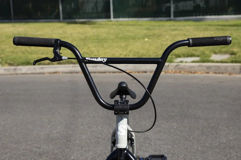 Vue rapprochée du guidon noir d'un vélo BMX Sunday sur une route asphaltée.