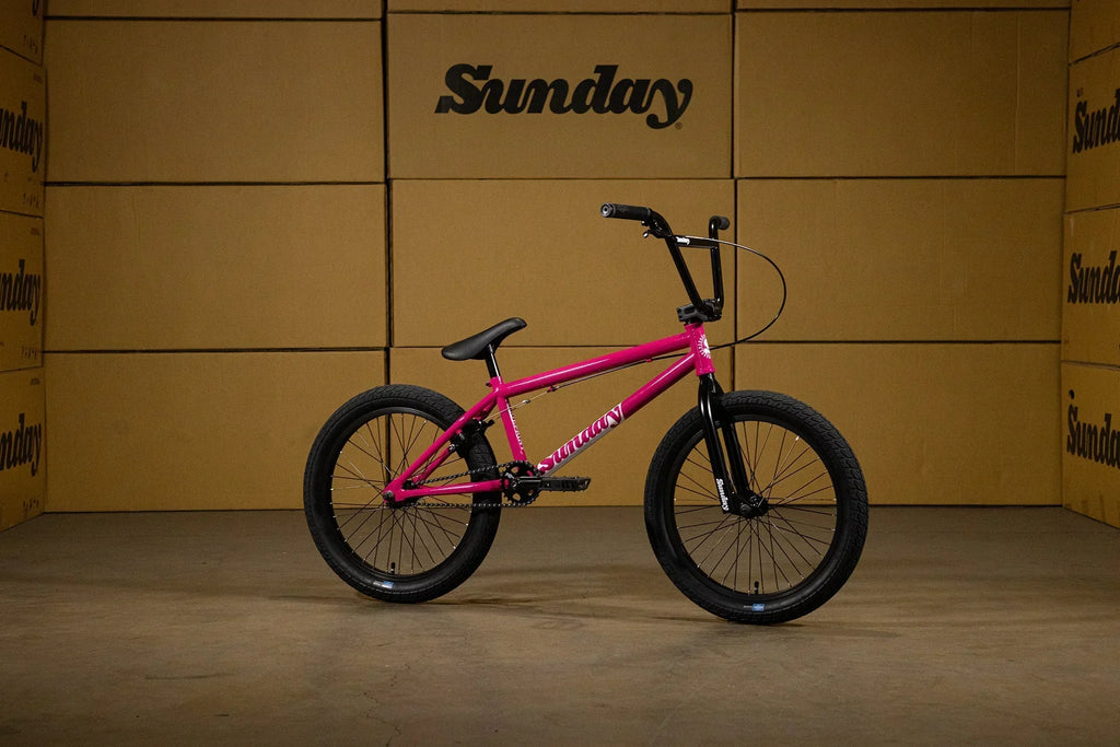 Vélo BMX Sunday rose vif avec pneus noirs sur fond de cartons de stockage.