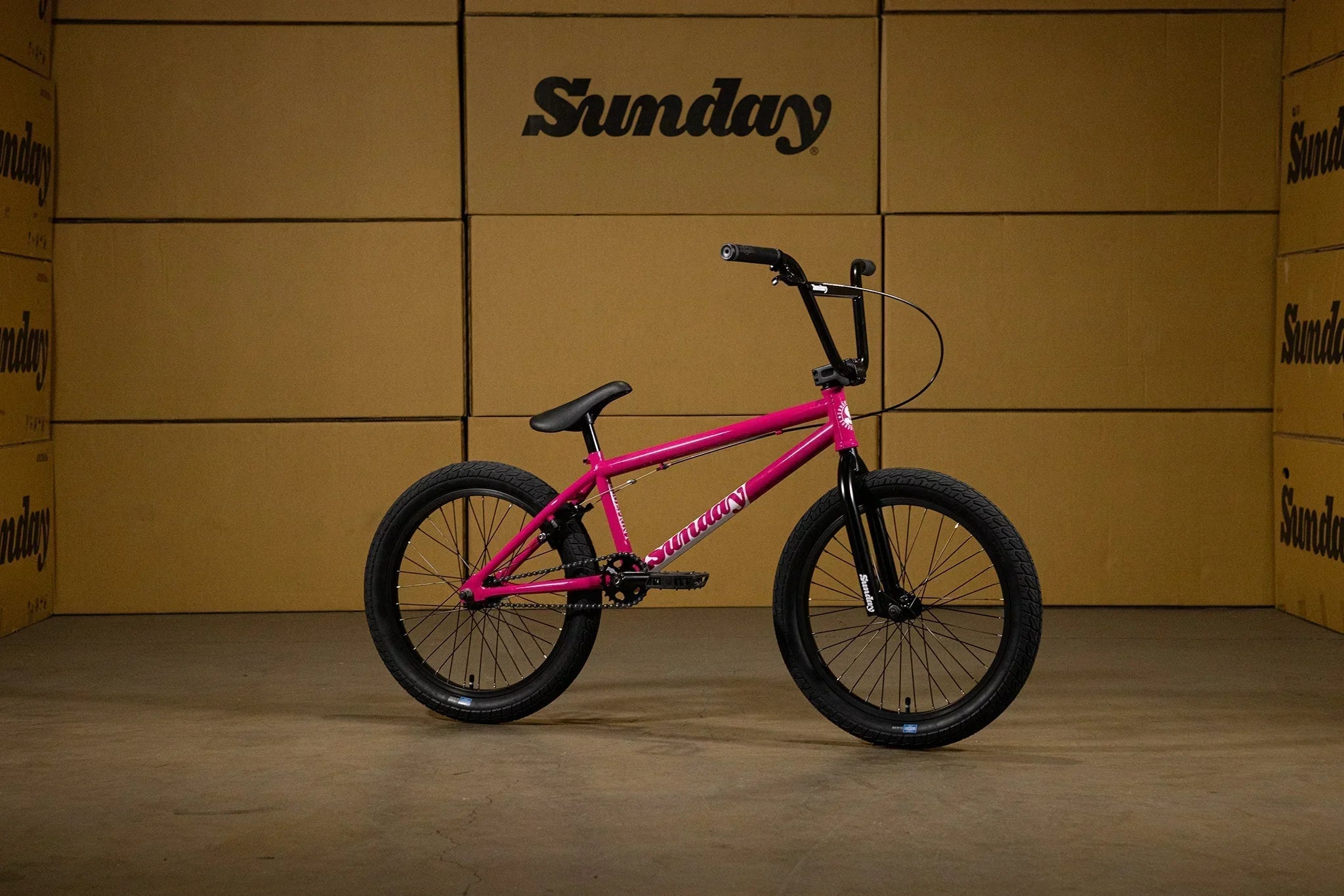 Vélo BMX Sunday rose vif avec pneus noirs sur fond de cartons de stockage.