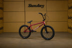 Vélo BMX Sunday rouge avec cadre métallique et pneus noirs sur fond de cartons.