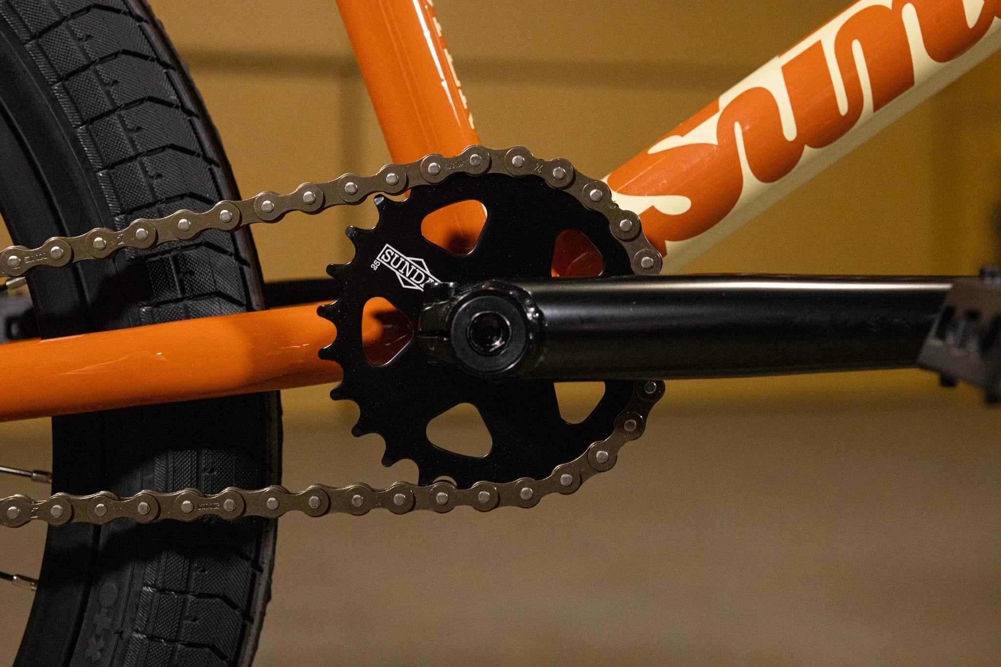 Gros plan du plateau Sundae noir et de la chaîne sur un cadre de vélo BMX orange.