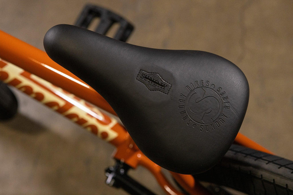 Gros plan d'une selle de vélo BMX noire avec logo embossé sur un cadre orange.