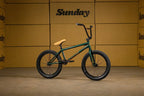 Vélo BMX vert avec selle beige et guidon noir sur fond de cartons Sunday