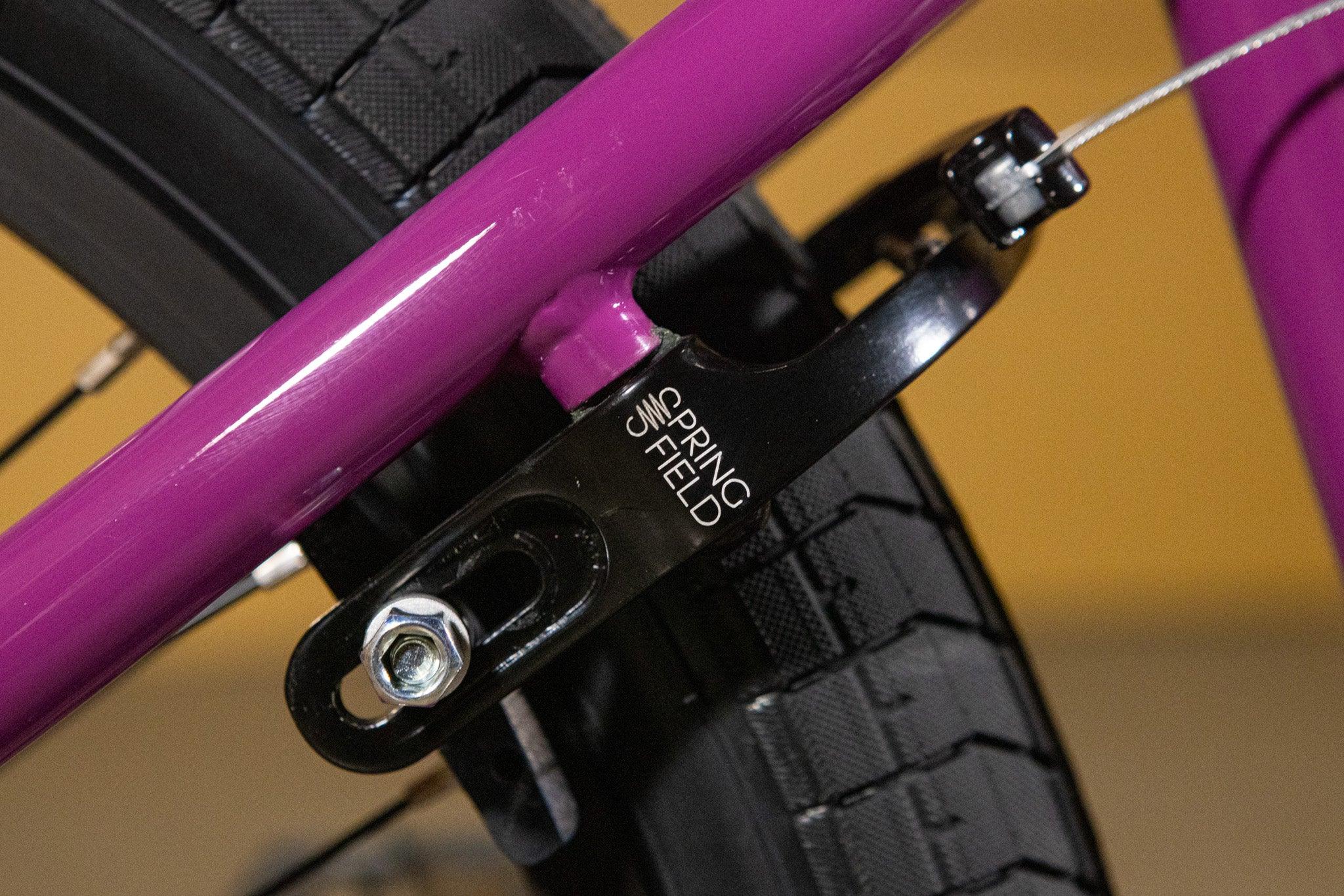 Détail du frein V-brake noir sur un cadre de vélo de couleur violette.