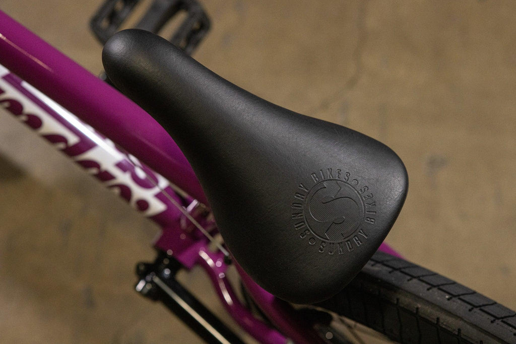 Gros plan d'une selle de vélo BMX noire avec logo embossé sur cadre violet.