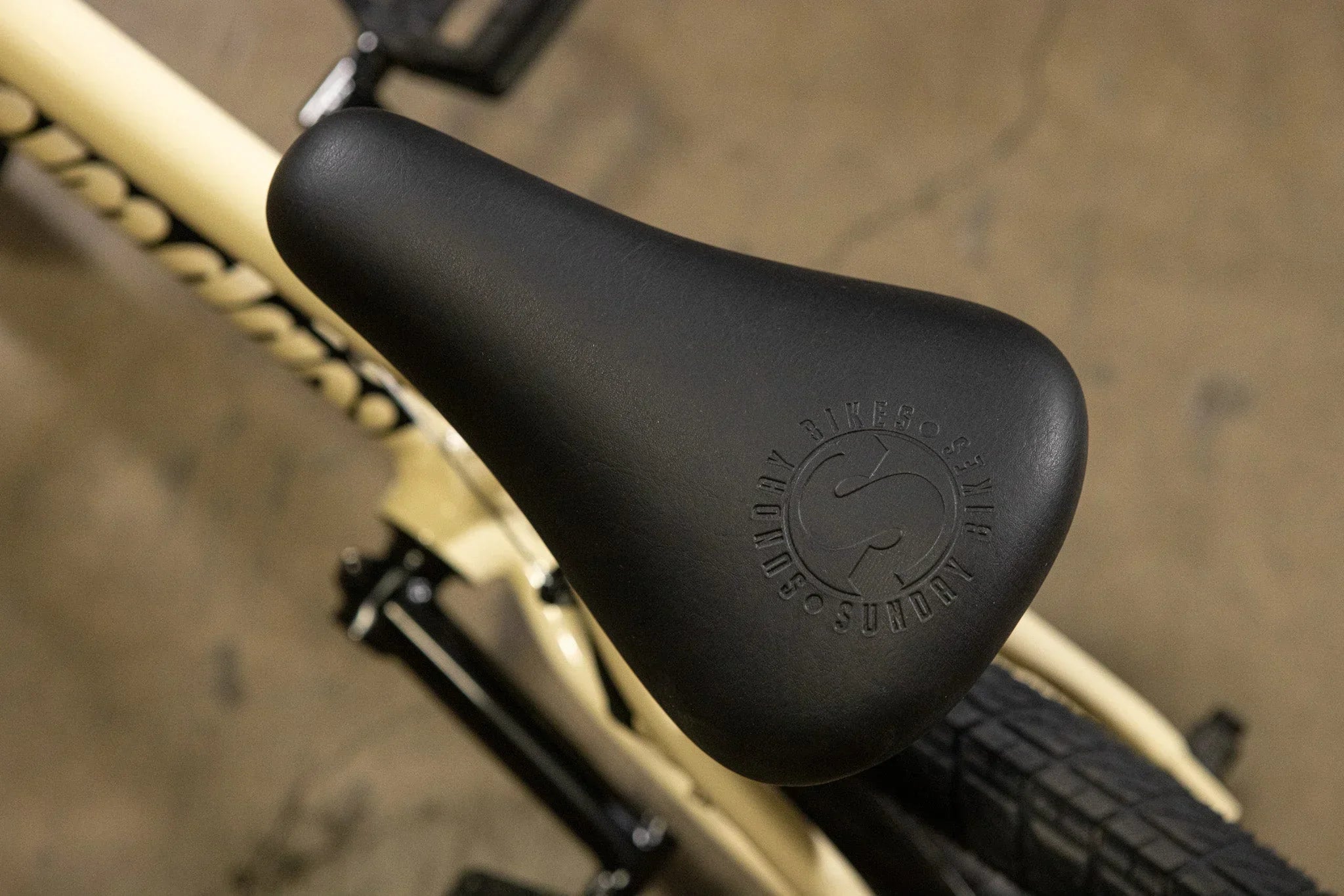 Gros plan d'une selle de vélo BMX noire avec logo Sundry Bikes sur un cadre beige.
