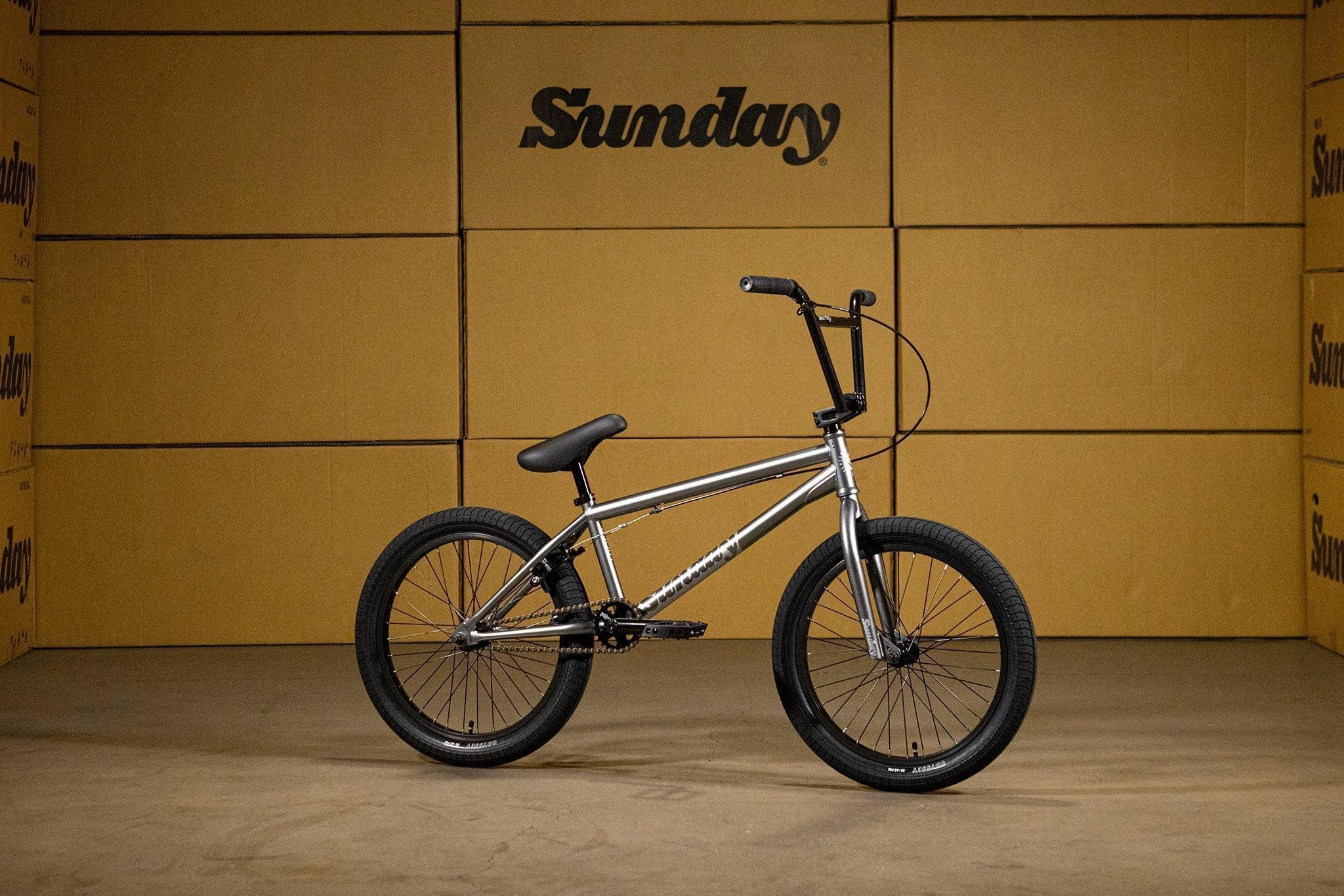 Vélo BMX argenté avec pneus noirs sur fond de cartons Sunday