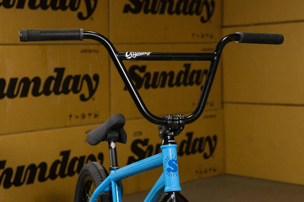 Guidon de vélo BMX noir avec poignées et cadre bleu devant des cartons Sunday