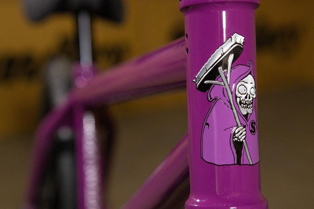 Gros plan d'un cadre de vélo BMX violet avec un autocollant de faucheuse stylisée.