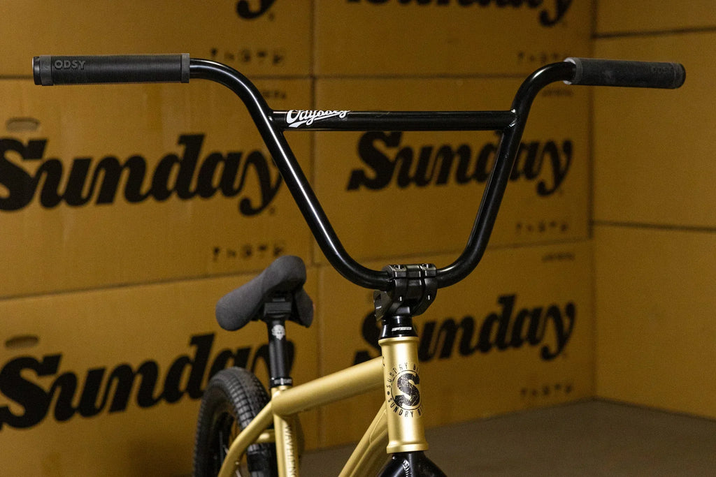 Guidon de BMX noir installé sur un cadre doré devant des boîtes Sunday