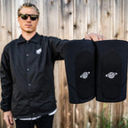 Person holding a pair of black BMX knee pads with white logo, outdoor wooden fence background.